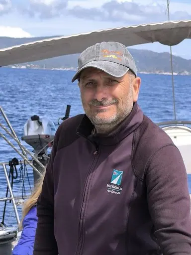 Un uomo con il berretto sorride a bordo, con la costa sullo sfondo