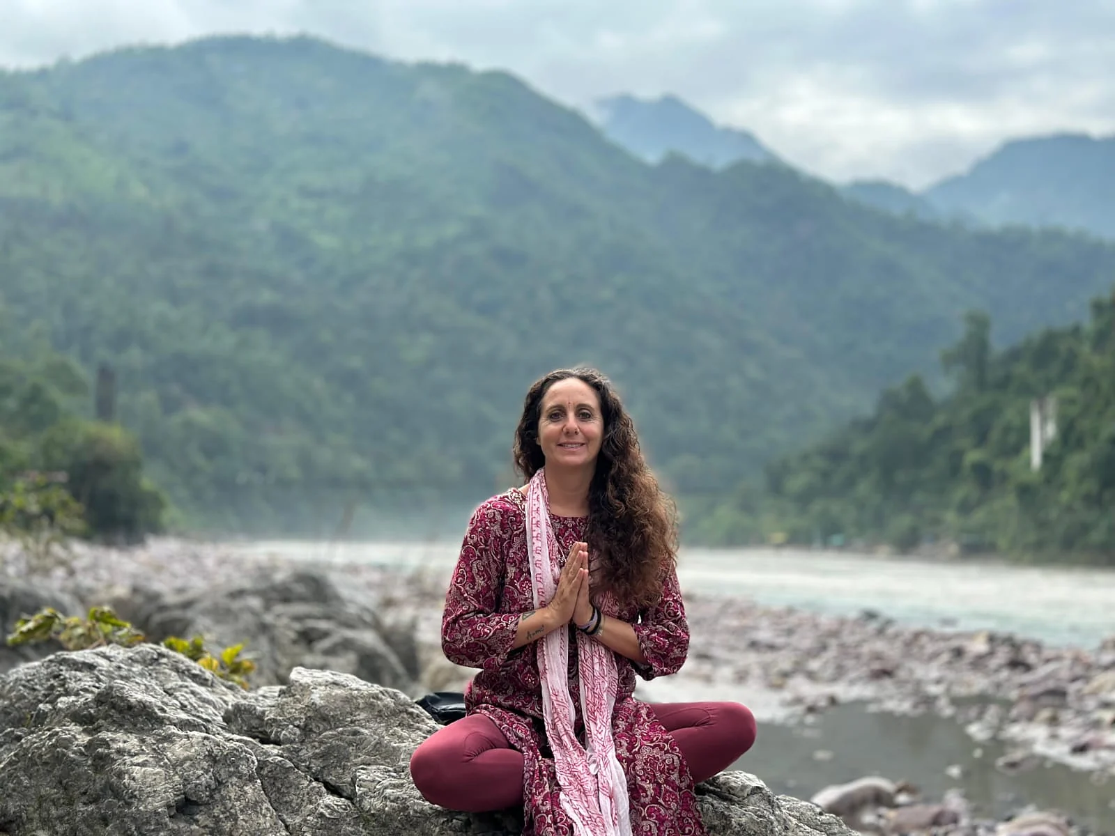 Laura Buoncristiani seduta a gambe incrociate su una roccia lungo il Gange, a mani giunte, con le montagne verdi alle spalle