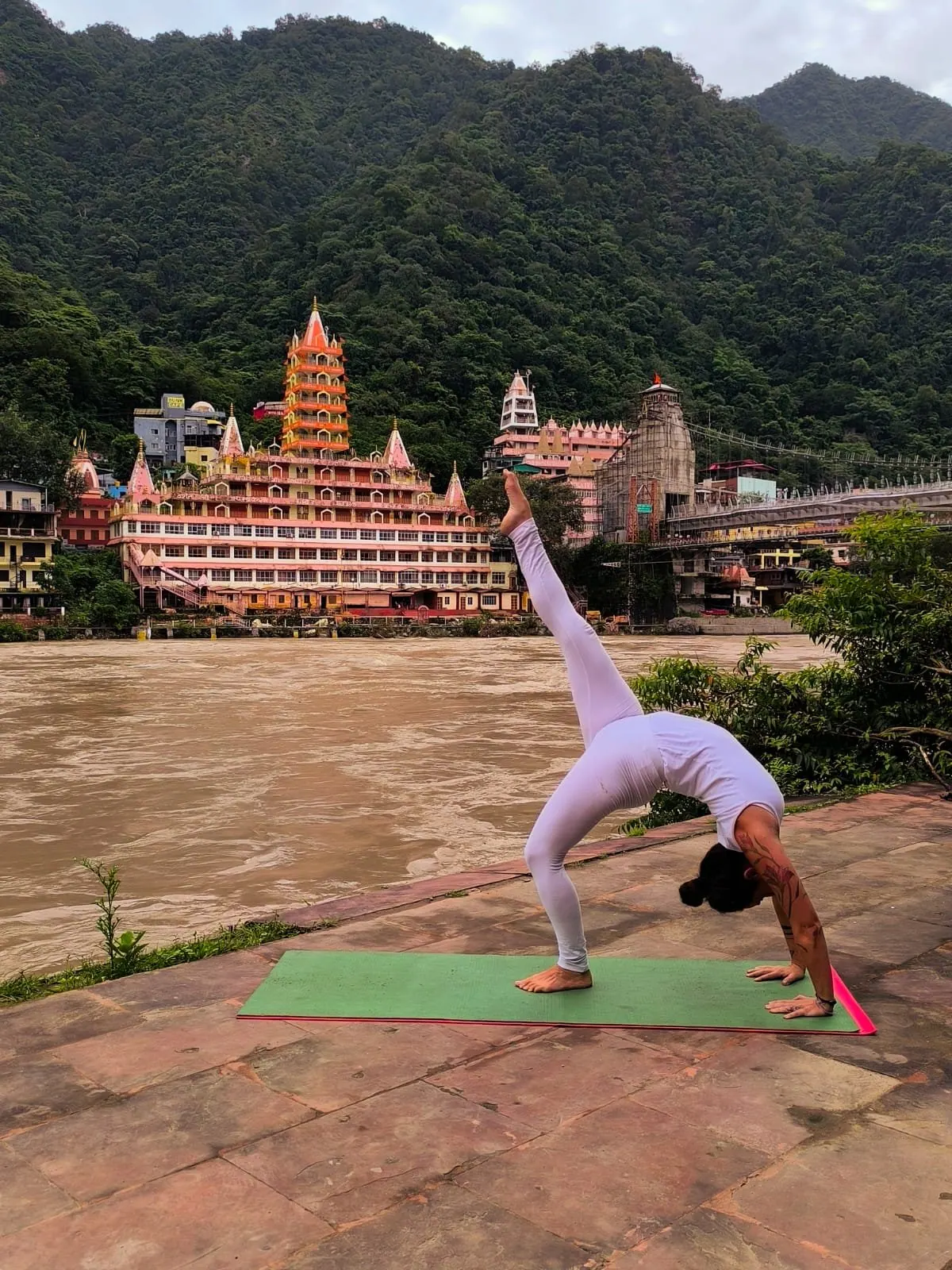 Laura in una posizione a ponte con una gamba tesa verso il cielo, sulla riva del Gange davanti ai templi di Rishikesh