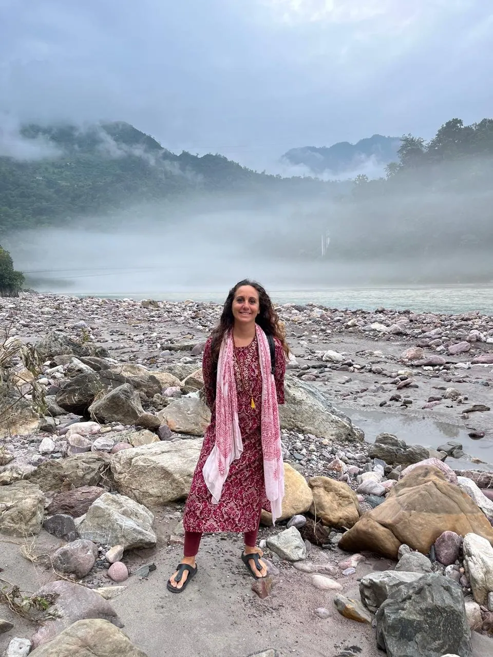 Laura in abito tradizionale rosso sulla riva sassosa di un fiume, fra le montagne avvolte dalla nebbia