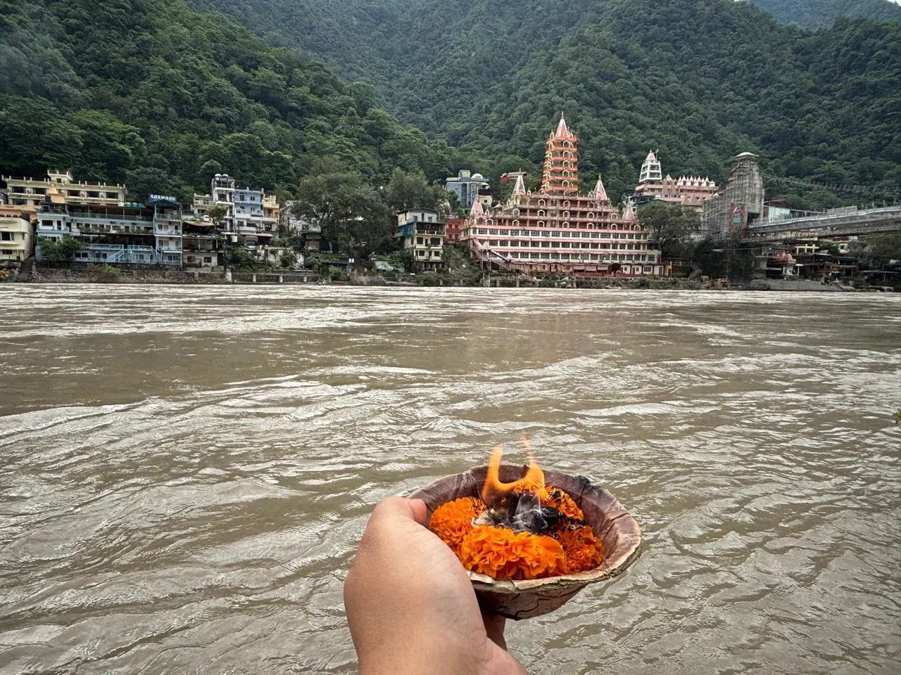 Una mano offre al Gange una coppa di foglie con fiori arancioni e una fiamma, davanti ai templi di Rishikesh