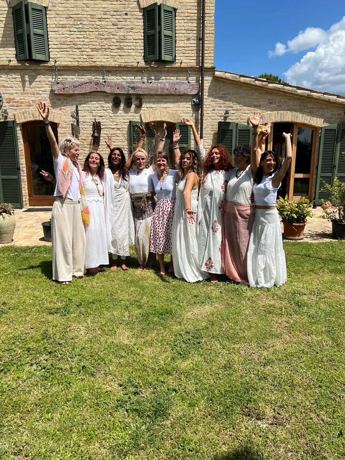 Donne vestite di bianco a braccia alzate sul prato davanti a un casale in pietra
